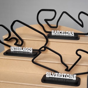 May include: Three black 3D printed race track models on a wooden shelf. The tracks are for Interlagos, Barcelona, and Silverstone. Each track has a white nameplate.