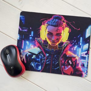 May include: Black mouse mat featuring a cyberpunk-style illustration of a person with yellow headphones, set against a neon cityscape. A red and black wireless mouse sits beside the mat. The person has blue hair and is wearing a black jacket.