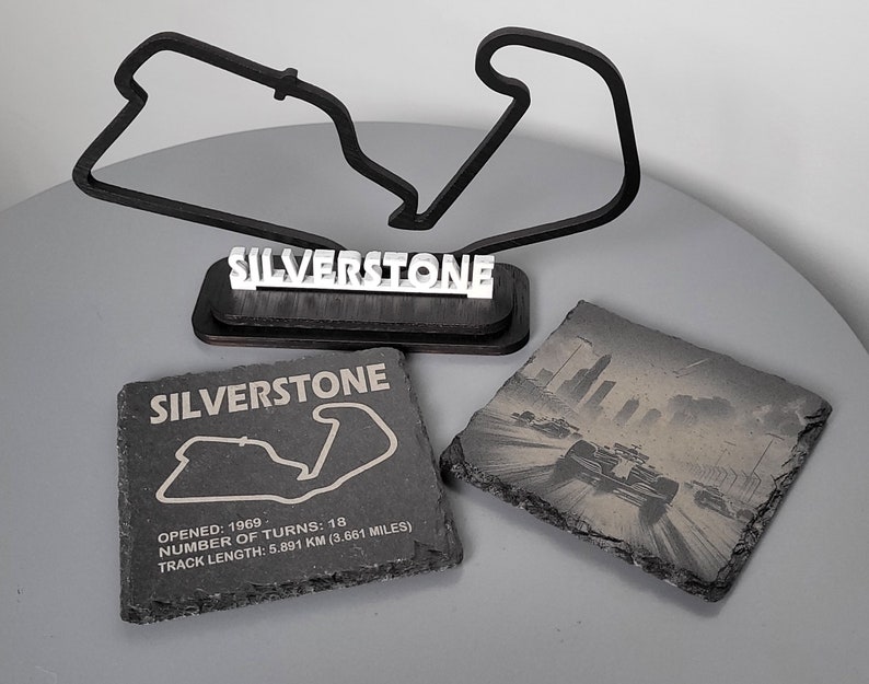 May include: Black and white image of the Silverstone race track on a slate coaster. The coaster has the words "SILVERSTONE" printed on it. The text below the track image reads "OPENED: 1969, NUMBER OF TURNS: 18, TRACK LENGTH: 5.891 KM (3.661 MILES)".