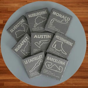 May include: Eight grey slate coasters with white text and track outlines. The coasters feature the names of famous race tracks, including Silverstone, Nurburgring, Monaco, Budapest, Austin, Hockenheimring, Le Castellet, and Barcelona. Each coaster includes the year the track opened, the number of turns, and the track length.