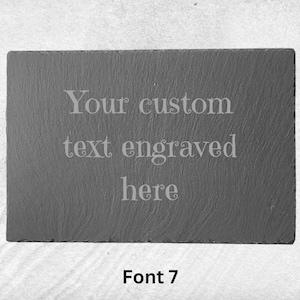Pode incluir: Uma placa de ardósia preta com o texto "Your custom text engraved here" em uma fonte decorativa. O texto é de cor cinza claro. A placa está sobre uma superfície de areia branca. O texto "Font 7" está abaixo da placa.