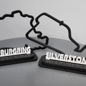 May include: Two black 3D printed race track outlines on wooden bases. The track on the left is labelled "N&uuml;rburgring" and the track on the right is labelled "Silverstone".