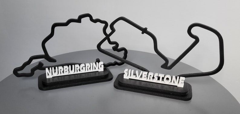 May include: Two black 3D printed race track outlines on wooden bases. The track on the left is labelled "N&uuml;rburgring" and the track on the right is labelled "Silverstone".