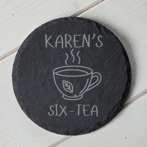 May include: A circular, dark grey slate coaster with the words "KAREN'S SIX-TEA" etched in light grey. A line drawing of a teacup with steam rising from it is in the center. The coaster is on a white wooden surface.