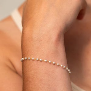 May include: A delicate bracelet featuring small, white pearls strung on a thin gold chain. The bracelet is worn on a wrist, showcasing its simple elegance. The pearls are evenly spaced, creating a classic and timeless look.