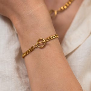 May include: Gold-toned bracelet with a toggle clasp, worn on a wrist. The bracelet features a curb chain design. A matching gold necklace is visible in the background. The person is wearing a white shirt.