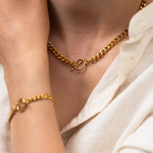 May include: A gold-colored chain necklace and bracelet set. The necklace and bracelet feature a curb chain design with a toggle clasp. The jewelry is worn against a white shirt.