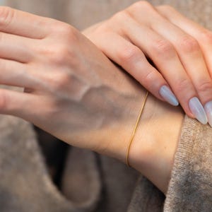 May include: A delicate gold bracelet is worn on a wrist. The thin, simple chain is a bright gold color. The person is wearing a neutral-colored sweater. The nails are painted a light blue color.