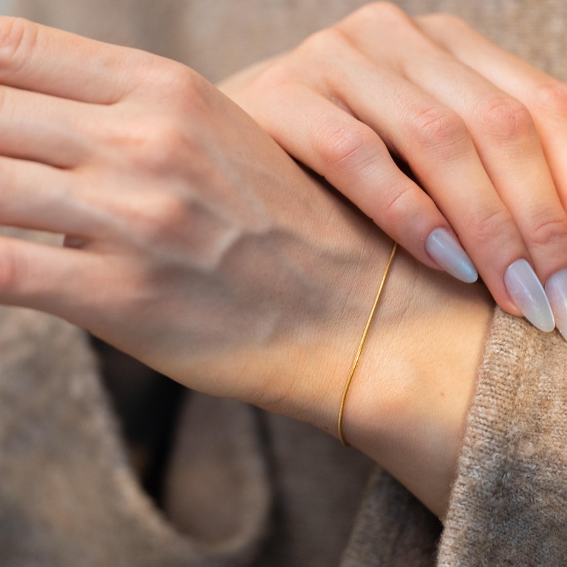 May include: A delicate gold bracelet is worn on a wrist. The thin, simple chain is a bright gold color. The person is wearing a neutral-colored sweater. The nails are painted a light blue color.