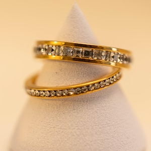 May include: Two gold rings, one with square-cut, the other with round-cut, clear gemstones. The rings are displayed on a white, cone-shaped stand. The top ring has square stones, the bottom has round stones. The rings are gold in colour.