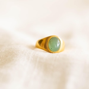 May include: A gold ring featuring a light green oval gemstone. The ring is set in a bezel and sits on a white, textured fabric. The band of the ring is thick and has a smooth, polished finish.