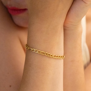 May include: A gold-colored chain bracelet with a classic curb link design. The bracelet is worn on a wrist, and the links are visible. The bracelet is a simple yet elegant piece of jewelry.