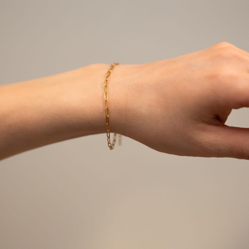 May include: A gold-colored chain bracelet with rectangular links is worn on a wrist. The bracelet is simple and elegant, with a delicate design. The background is a solid, neutral color.