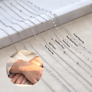 May include: A collection of silver-toned metal chains including a large paperclip, pearl bead, twist, satellite, ball, and tiny paperclip chain. A circular inset shows a wrist wearing a silver-toned chain bracelet. Text labels identify each chain style.