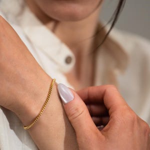 May include: A delicate gold chain bracelet with a square link design. The bracelet is worn on a wrist, and the gold color contrasts with the skin tone. The bracelet is a simple yet elegant piece of jewelry.