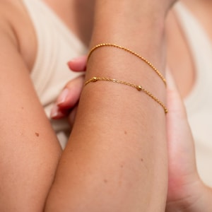 May include: Two gold-toned bracelets worn on the wrist. The top bracelet is a simple, thin chain. The bottom bracelet is a delicate chain featuring small, evenly spaced gold spheres along its length, creating a subtle and refined appearance.