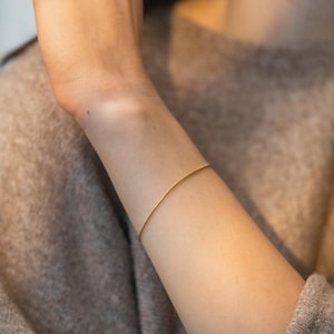 May include: A thin, minimalist gold-toned wire bracelet encircling a wrist. The bracelet features a simple, smooth, and continuous circular design with a polished metallic finish that reflects light, creating a subtle and understated appearance.