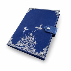 May include: A deep blue leather-textured journal featuring a silver embossed illustration of a castle and two flying dragons. The cover is accented with ornate silver metal corner protectors and a matching strap with a silver snap closure.