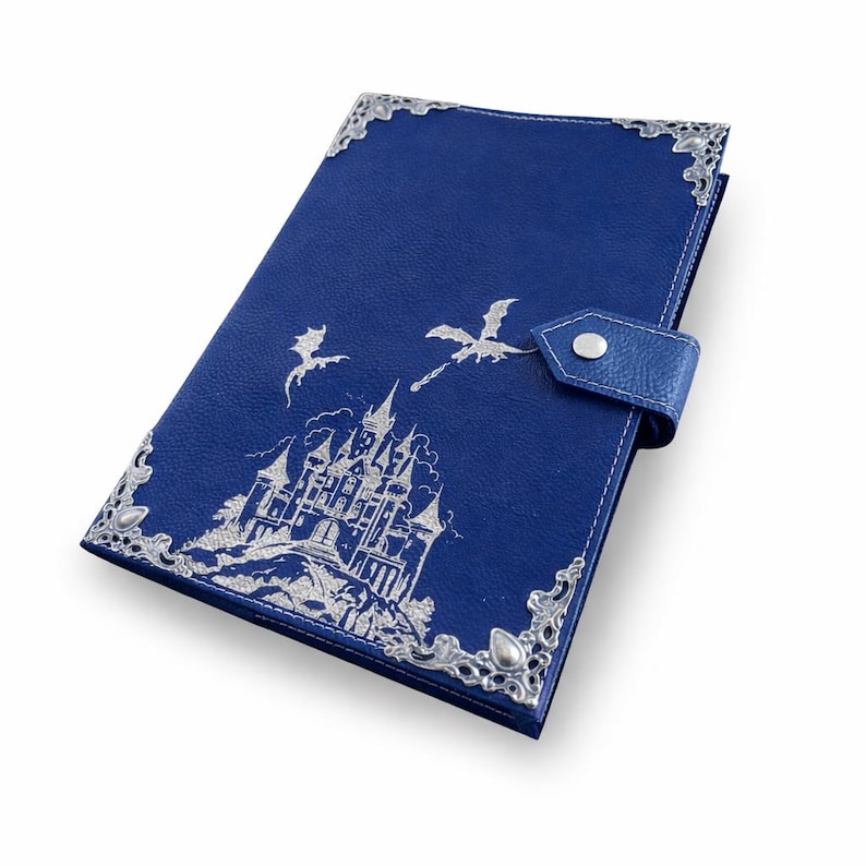 May include: A deep blue leather-textured journal featuring a silver embossed illustration of a castle and two flying dragons. The cover is accented with ornate silver metal corner protectors and a matching strap with a silver snap closure.