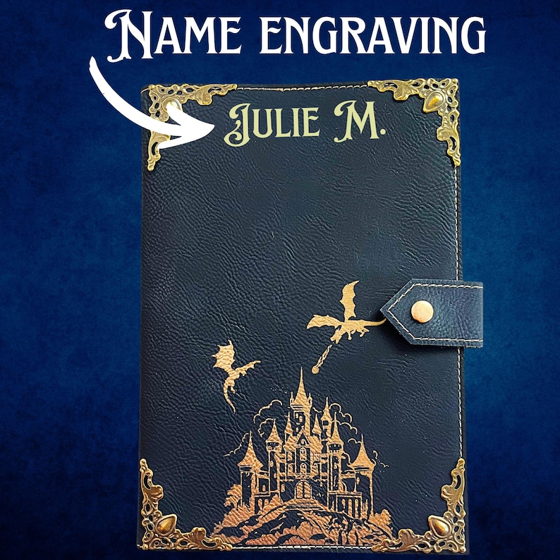 May include: A black leather-textured journal cover featuring gold-colored metal corner protectors, a snap closure, and a gold-colored graphic of a castle with two flying dragons. The name JULIE M. is engraved in gold lettering at the top.