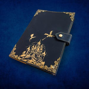 May include: A black leather journal with gold accents. The cover features a gold illustration of a castle and dragons. Gold corner protectors and a snap closure complete the design. The journal is a fantasy-themed item.