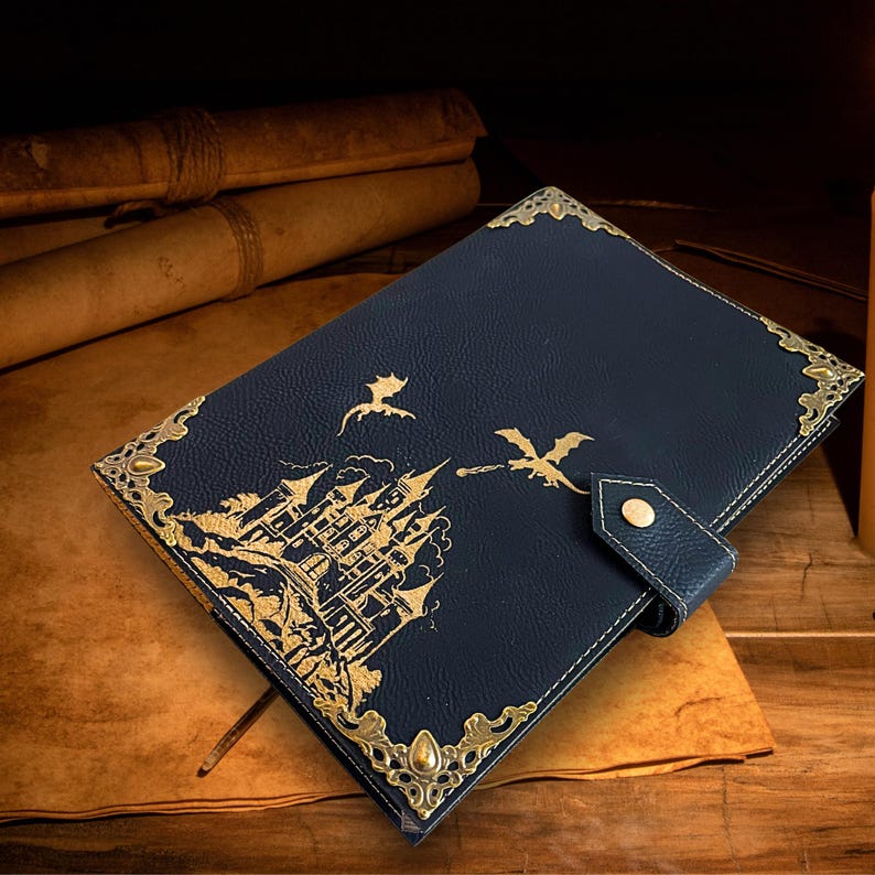 May include: A navy blue leather journal with a gold castle and dragon design. The journal features gold corner accents and a snap closure. The cover art depicts a castle with dragons flying above it, creating a fantasy theme.
