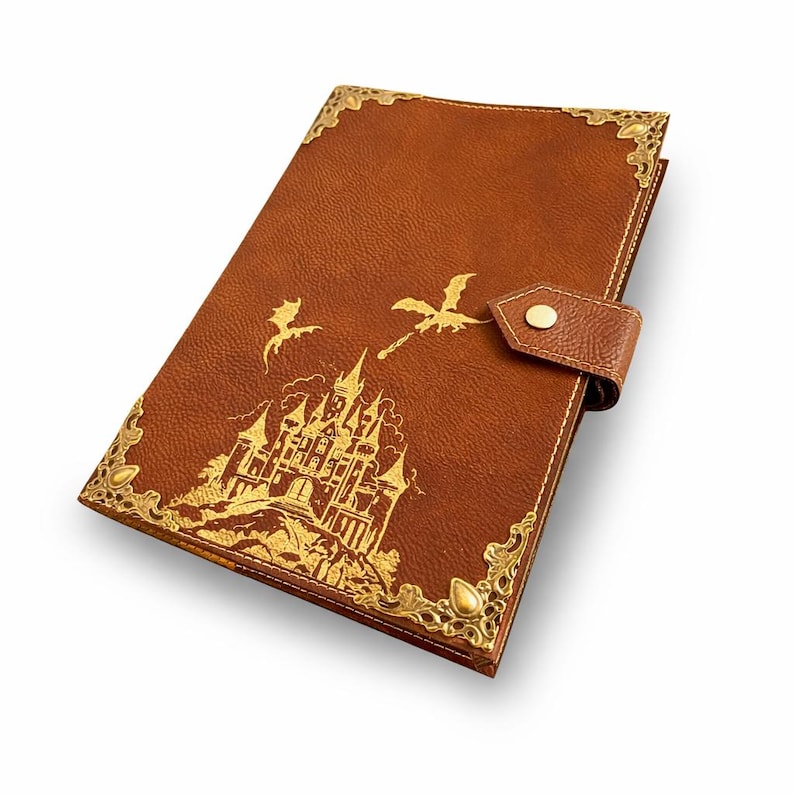 May include: A brown leather-textured journal featuring a gold-colored embossed castle and two flying dragons on the front cover. The notebook includes ornate gold metal corner protectors and a matching strap closure with a single gold snap button.
