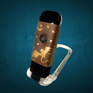 May include: A brown and black vape pen with a gold dragon and stars design. The pen is on a clear plastic stand.