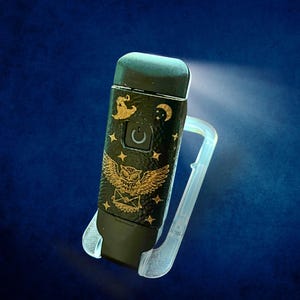 May include: Black and gold decorative lighter with an owl and celestial design. The lighter has a power button and is held in a clear plastic holder. The design includes a witch hat, moon, stars, and an owl holding a letter.