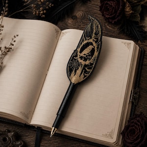 May include: A black pen with a feather-shaped top featuring a gold raven design, spiderwebs, and bats. The pen rests on an open, lined journal with decorative corner designs. The pen's design is reminiscent of gothic or fantasy themes.
