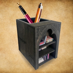May include: A dark stained wooden desk organizer with a pen holder and small shelves. The pen holder contains a pink and a gold pen. The shelves hold small books and notebooks. The organizer has decorative carved details.