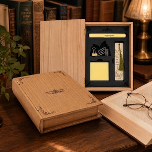 May include: A wooden book-shaped box with a hinged lid, containing stationery items. Inside, a highlighter, paper clips, a sticky note pad, and a decorative bookmark are visible. The box is open, revealing the contents, and sits on a wooden surface.