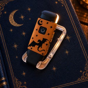 May include: A portable orange and black electronic lighter featuring a dragon silhouette, crescent moon, and star pattern. The device has a central power button with a power symbol and is attached to a clear, translucent plastic carabiner clip.