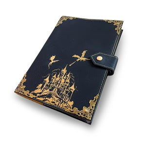 May include: A dark blue journal with gold accents and a decorative clasp. The cover features a gold illustration of a castle, dragons, and ornate corner detailing. The journal is closed with a leather strap and a gold button.