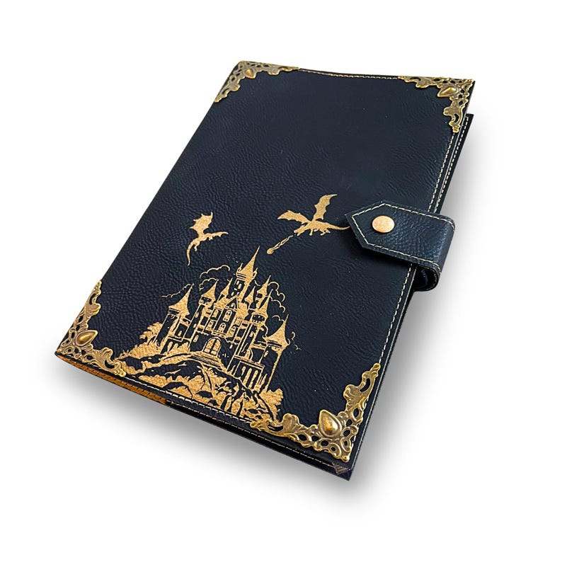 May include: A dark blue journal with gold accents and a decorative clasp. The cover features a gold illustration of a castle, dragons, and ornate corner detailing. The journal is closed with a leather strap and a gold button.