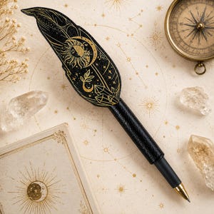 May include: A black feather-shaped pen featuring gold foil illustrations of a sun, moon, stars, and crystals. The pen has a textured black barrel and a metallic gold tip, designed with a detailed celestial motif across the top surface.
