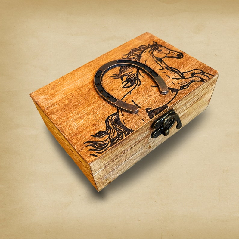 Pu&ograve; includere: Una scatola di legno con un design a forma di cavallo e ferro di cavallo. La scatola ha un coperchio con cerniera e una chiusura in metallo.