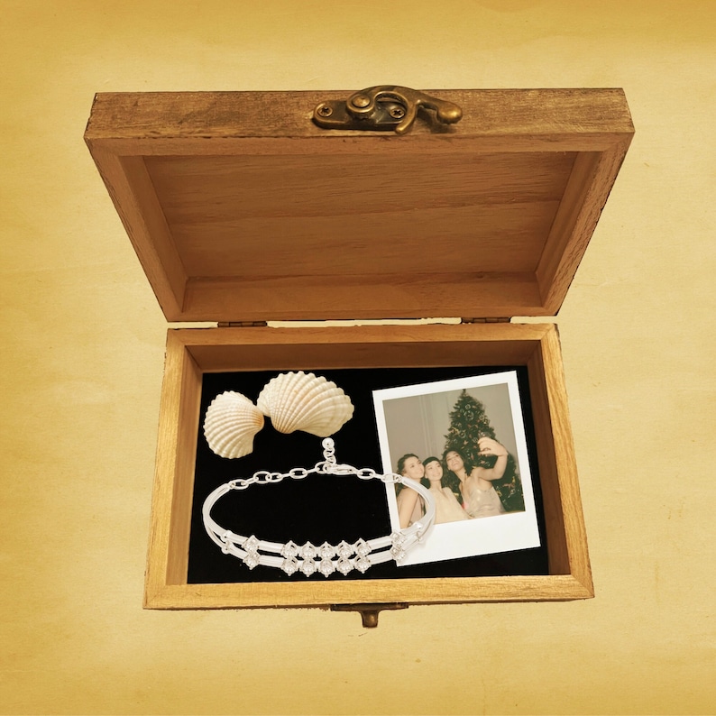 Pu&ograve; includere: Una scatola di legno con un coperchio incernierato, aperta per rivelare una conchiglia, un braccialetto d'argento e una fotografia incorniciata di quattro persone in piedi davanti a un albero di Natale.