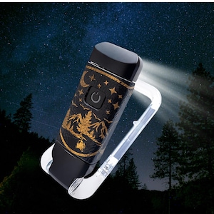 May include: A portable black flashlight with a gold-colored graphic print featuring a crescent moon, stars, pine trees, and a mountain range. The device includes a power button and a clear, transparent carabiner clip attached to the base.