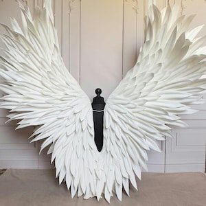 May include: A pair of large, white, feathered angel wings. The wings are made of many individual feathers that are layered and overlapping. The wings are attached to a black stand.