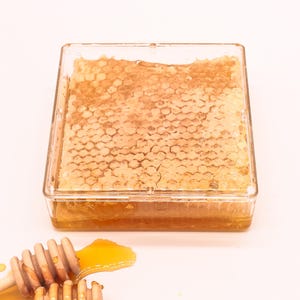May include: A clear square container filled with honeycomb, a natural honey product. Two wooden honey dippers are on a white surface with a small amount of honey spilled around them.