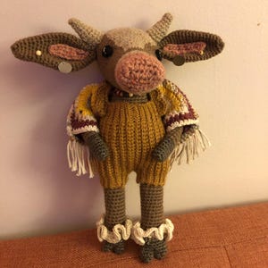 May include: A handmade crochet cow doll with a mustard yellow jumpsuit, brown ears, and a pink nose. The doll has decorative fringe on the sleeves and white frills around the ankles. The cow has small horns and a beaded necklace.