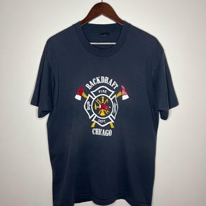 May include: A dark blue t-shirt with a white graphic of a firefighter's emblem. The emblem features a crossed axe, a fire hydrant, and the words "Backdraft Fire Dept. Chicago".