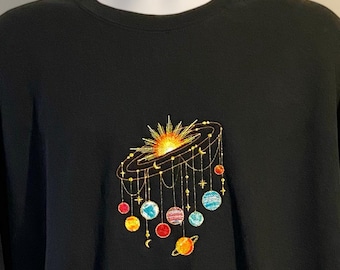 Könnte beinhalten: Ein schwarzes Langarm-Sweatshirt mit einer zentralen Stickerei, die eine leuchtende Sonne und verschiedene bunte Planeten zeigt, welche an feinen Linien hängen und ein mobileartiges Arrangement auf der Brust bilden.