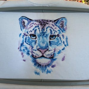 May include: Embroidered white sweatshirt featuring a detailed snow leopard face in shades of blue, purple, and pink. The design is framed by a gray embroidery hoop, measuring 28 x 20 cm (11.8" x 7.9").