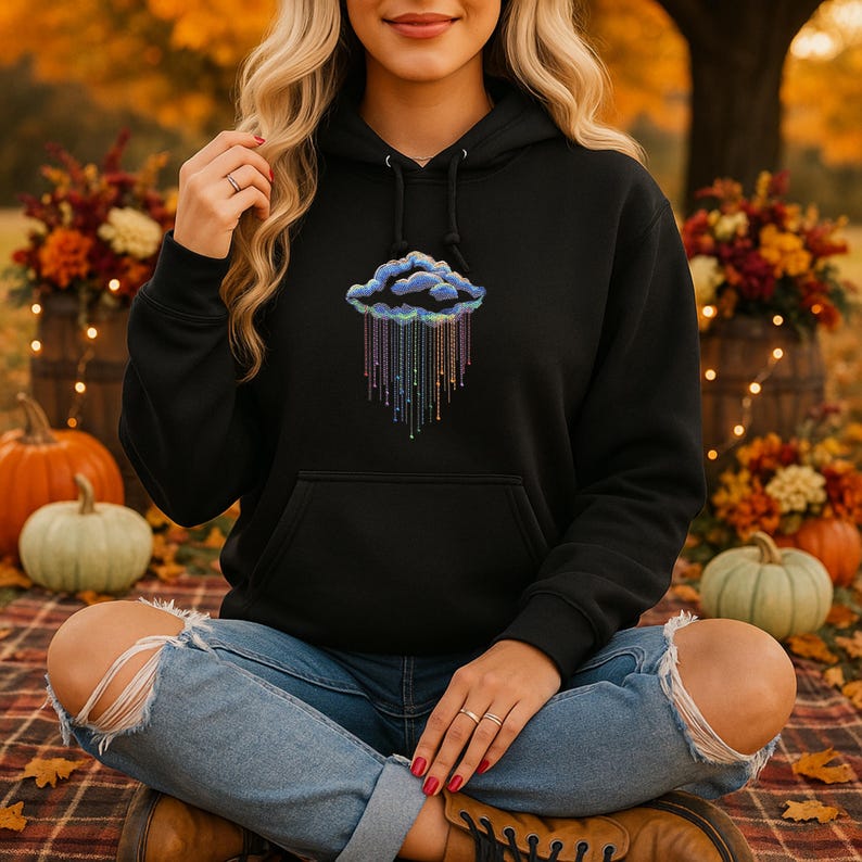 May include: Black hoodie with a cloud design and rainbow rain. The cloud is outlined in blue and the rain is a gradient of colours. The sweatshirt has a front pocket and a drawstring hood.