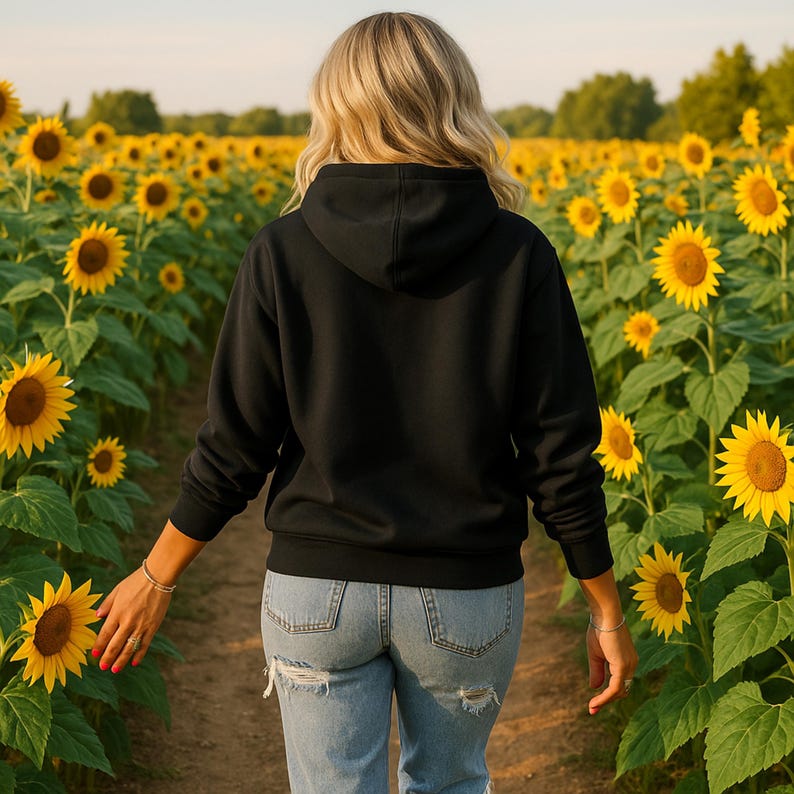 May include: A person wearing a black hooded sweatshirt and blue jeans walks through a field of sunflowers. The sunflowers are in full bloom, with bright yellow petals and dark brown centres. The person is blonde and is walking away.