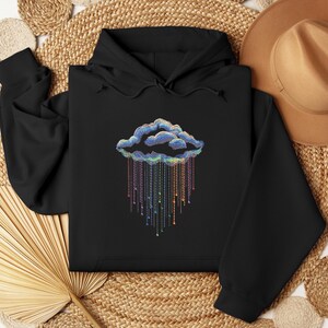 May include: Black hoodie with a cloud design and colourful rain. The cloud is outlined in blue and the rain is made of colourful strings with small beads. The sweatshirt is made of a soft material.