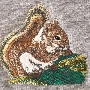 May include: An embroidered patch on a heather gray fabric featuring a brown and white squirrel with a bushy tail, sitting on a patch of textured green grass. The squirrel is depicted in profile, showcasing detailed stitching in shades of brown.