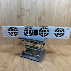 May include: A white electronic device with four circular fan openings and a black power switch. The device is mounted on a silver scissor lift. The background is a light brown wooden surface.